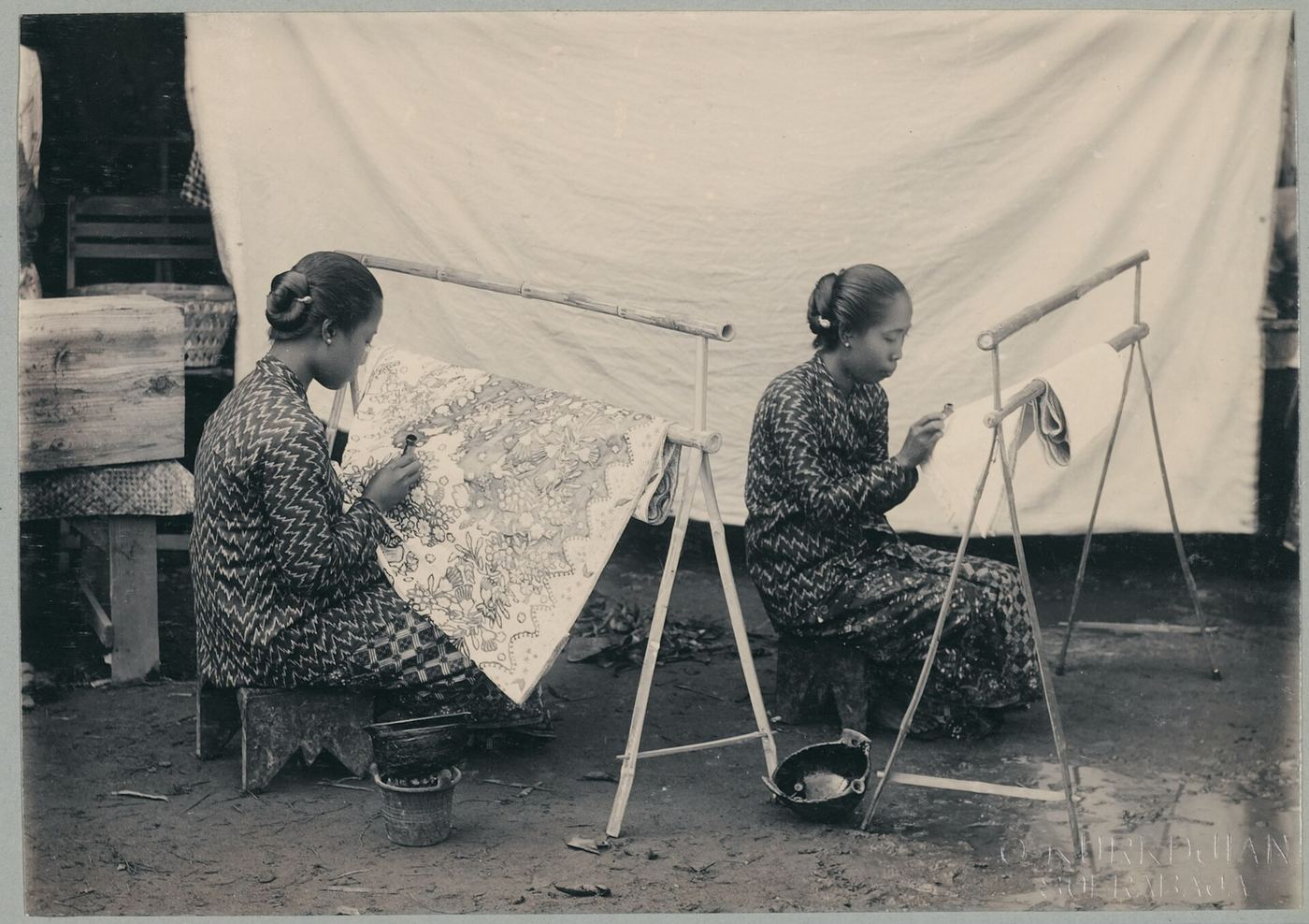 Dua perajin wanita menenarkan kain batik di atas rangka kayu pada pasar malam Surabaya, 1905